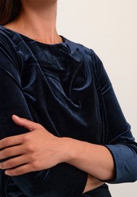 Mörkblå sammetstopp med en draperad halsringning, har långa ärmar och en mjuk textur, som visar mjuka veck och en formsydd design.