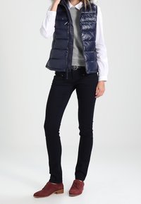 Chaleco tipo puffer de color navy con textura acolchada, llevado sobre un suéter gris y una camisa blanca. Combinado con jeans oscuros y zapatos de cordones burdeos.