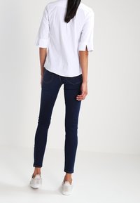 Biała koszula z kołnierzykiem i rękawami za łokcie, w połączeniu z ciemnoniebieskimi obcisłymi jeansami i jasnymi sneakersami, widziana od tyłu.