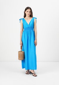 Blå maxi kjole med V-hals, smocket talje og bindesløjfer. Kombination med en vævet håndtaske og sorte flip-flops. Glat tekstur, afslappet design.