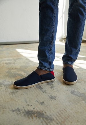 Person wearing dark blue slip-on shoes with red heel accents and beige soles, paired with blue jeans, standing on a textured floor.