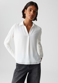 Weiße Bluse mit langen Ärmeln, kragenlos und leicht mit V-Ausschnitt. Glatte Stoffqualität, lockere Passform, kombiniert mit dunklen Jeans.