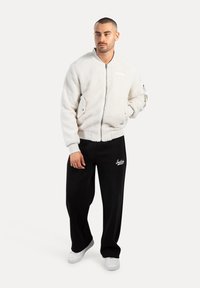 Hombre vistiendo una chaqueta bomber de forro polar blanca, pantalones de chándal negros y zapatillas blancas, de pie contra un fondo liso.