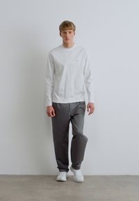 Fehér hosszú ujjú ing kis logóval, szürke nadrággal és fehér sportcipővel párosítva. Az anyagok puhanak tűnnek, laza szabással.