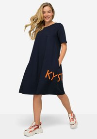 Navy blue short-sleeve dress with a loose fit, featuring large orange lettering "KY" on the front and side pockets, paired with chunky sneakers.