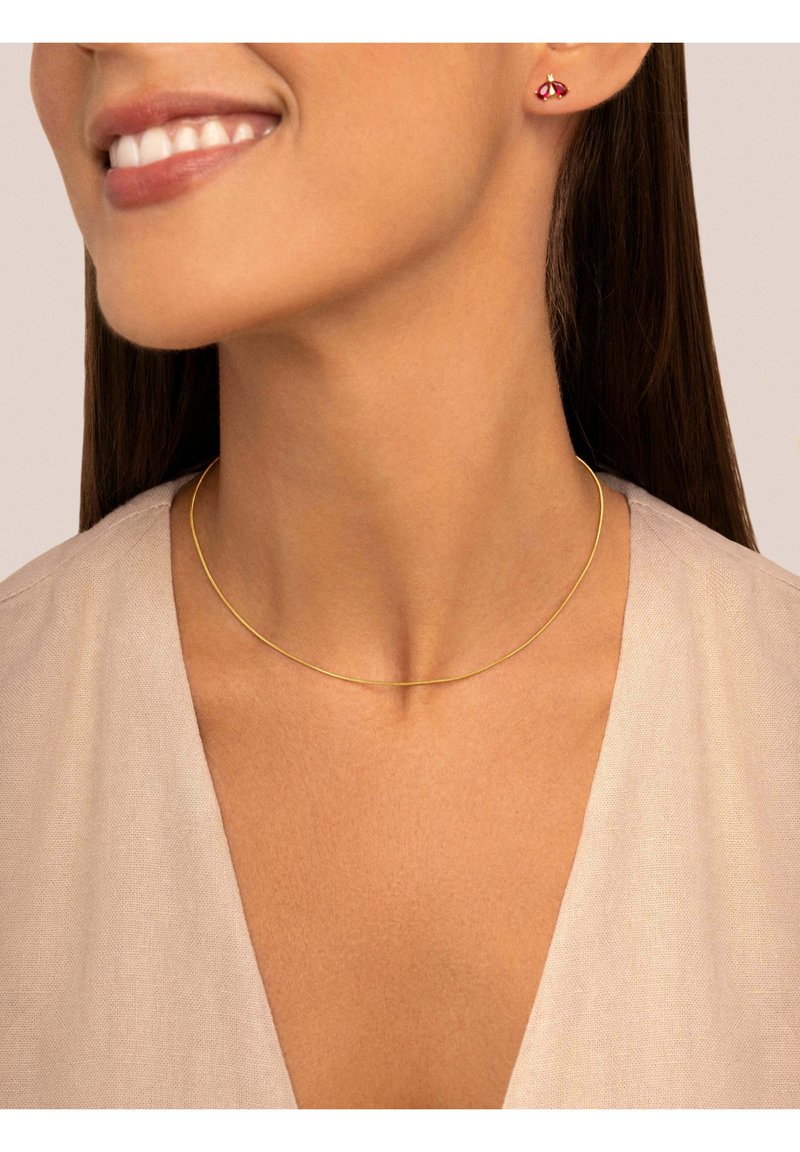 Collier ras du cou en ton doré avec un design élégant et fin. Le modèle porte des boucles d'oreilles en forme de clou rouges. Un vêtement beige clair et texturé est visible en arrière-plan.