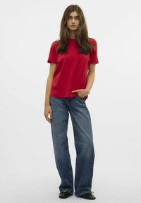 Rode katoenen t-shirt met korte mouwen, gecombineerd met lichtblauwe wijde denim jeans. Model staat met één hand in de zak, op een neutrale achtergrond.