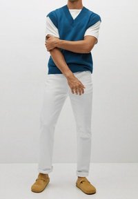 Modrý pletený svetr přes bílý tričko, doplněný bílými rovnými kalhotami a hnědými mokasíny. Hladké textury a jednoduchý design.