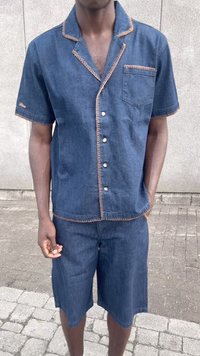 Mann iført blå denim kortermet skjorte med knapper og matchende shorts med beige sømmer, stående på et asfaltert underlag nær en grå vegg.