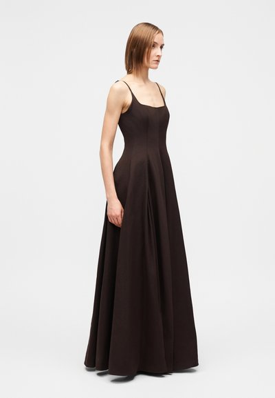 Mulher vestindo um vestido preto sem mangas, comprido até ao chão, com alças finas, parada de perfil contra um fundo claro simples.