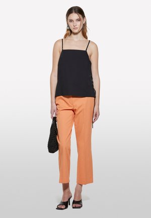 Camisole nera con spalline sottili e dettaglio di lacci laterali, abbinata a pantaloni arancioni a gamba dritta e mules nere con tacco.