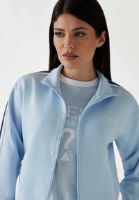 Lichtblauwe zip-up jack met een hoge kraag, voorzien van een wit "GUESS" logo en contrasterende accenten langs de mouwen. Gelaagd over een blauwe graphic t-shirt.