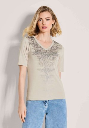 Beiges V-Ausschnitt-T-Shirt mit kurzen Ärmeln, verziert mit einem strukturierten Paisley-Muster in einem helleren Farbton, kombiniert mit hellblauen Jeans.