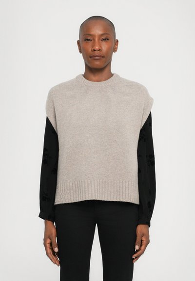 Maglione beige a maglia con orlo a coste e maniche corte, indossato sopra una camicia a maniche lunghe con motivo floreale nero.