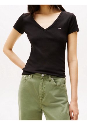 Vrouw draagt een nauwsluitend zwart T-shirt met V-hals en korte mouwen en olijfgroene broek, staand met één hand op haar heup.