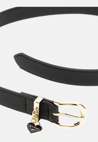 Curea din piele neagră cu o cataramă aurie strălucitoare, având un pandantiv în formă de inimă care spune "GUESS girl". Textură netedă și design elegant.