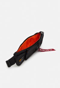 Fekete deréktáska narancssárga béléssel, több zsebbel, cipzáros záródással és piros "REMOVE BEFORE FLIGHT" címkével.