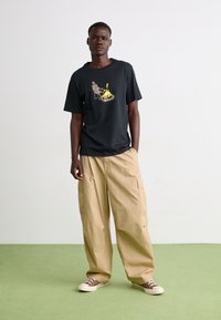 Una camiseta negra de manga corta presenta un gráfico de un esqueleto al lado de una fogata. Combinada con pantalones cargo beige claros y zapatillas casuales marrones.
