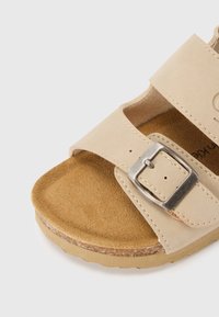 Μπεζ σανδάλι με δύο φαρδιές λουρίδες με μεταλλικό πόρπη, μαλακό σουέντ πάτο και ανθεκτική σόλα με αντίθετη υφή.