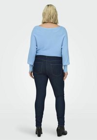 Kvinna med blont hår som bär en ljusblå ribbad tröja, mörka skinny jeans och svarta ankelboots, vänd bort från kameran mot en neutral bakgrund.