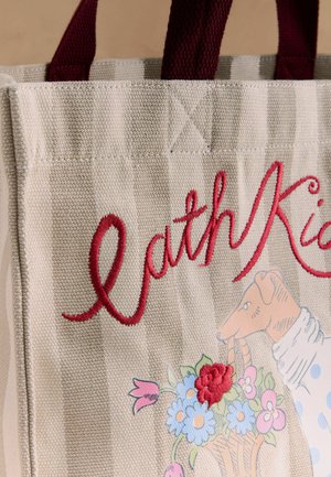 Csíkos bézs színű bevásárlótáska bordó fogantyúkkal, piros "Cath Kid" hímzett felirattal és egy kutyát ábrázoló illusztrációval, aki egy virágkosarat tart.