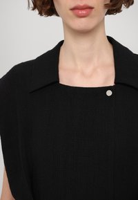 Fekete textúrált ruházat, állógallérral és diszkrét ezüstgomb részlettel. Széles, laza szabású és négyzet alakú dekoltázs jellemzi.