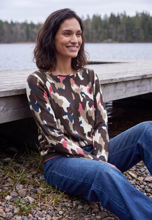 Mujer sonriente con cabello oscuro sentada en terreno rocoso junto a un muelle de madera, con una camisa de manga larga con patrón y vaqueros azules cerca de un lago.