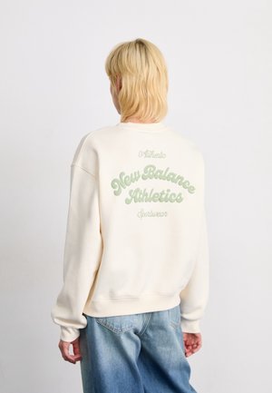 Personne aux cheveux blonds courts portant un sweat crème avec le texte vert "New Balance Athletics" dans le dos et un jean bleu ample.