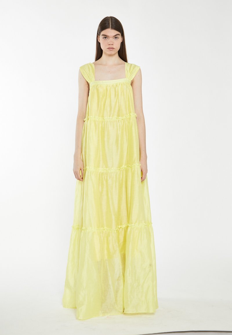 Junge Frau mit langen dunklen Haaren, die ein ärmelloses, hellgelbes, mehrlagiges Maxi-Kleid trägt und vor einem schlichten weißen Hintergrund steht.