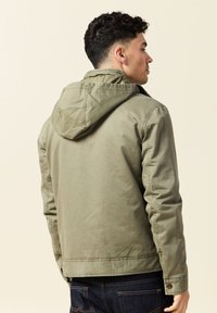 Man met zijn rug naar de camera, gekleed in een olijfgroen hoodiejack en donkerblauwe jeans, tegen een neutrale achtergrond.