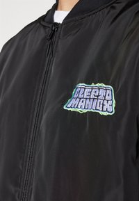 Črni bomberska jakna z zadrgo spredaj in barvno vezeno logotipno obližico na levem prsnem predelu. Gladek, sijoč teksturiran material.