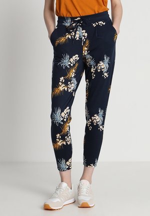 Osoba ubrana w granatowe spodnie w kwiatowy wzór z paskiem ze sznurkiem, pomarańczową koszulę oraz białe trampki, stojąca z rękami w kieszeniach.