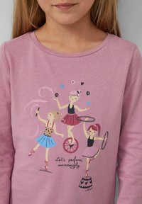 Rosa Langarmshirt mit Grafik von drei Künstlern: einer mit einem Einrad, einer mit Hulahoops und einer mit einem Stock, umgeben von verspielten Akzenten.