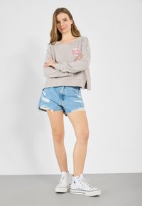 Lys grå avkortet hettegenser med rosa grafisk tekst, kombinert med slitte blå denimshorts. Modellen har på seg hvite høye sneakers.