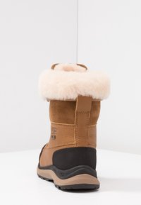 Botte d'hiver en daim marron et cuir avec un col en shearling crème duveteux, une semelle en caoutchouc noire, et des détails cousus au talon et sur les côtés.