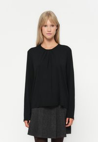 T-shirt noir à manches longues avec un col rond, présentant des plis discrets à l'avant. Associé à une jupe grise à carreaux, en tissu texturé.