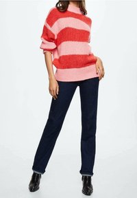 Kvinde iført en rød og pink stribet sweater, mørkeblå jeans og sorte ankelstøvler, der står med den ene hånd i lommen.