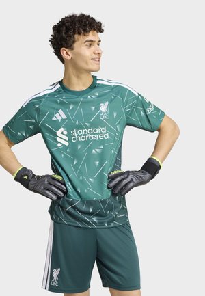 Joven con el uniforme verde y estampado de portero del Liverpool FC con guantes negros Adidas, manos en las caderas, mirando hacia un lado, sonriendo.