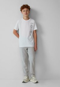 Witte katoenen T-shirt met bedrukte graphics aan de linkerkant, gecombineerd met lichtgrijze sweatpants en lichte sneakers.