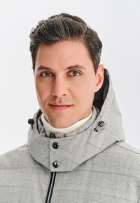 Jasnoszary płaszcz w wzory z wysokim kołnierzem, ozdobiony czarnymi guzikami i sznurkiem, noszony na białym golfie. Gładka tekstura.