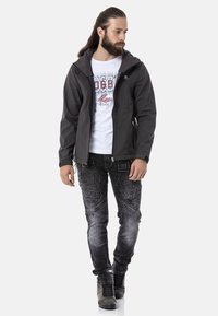 Schwarze Softshelljacke mit einem Reißverschluss vorne, grauem Kapuzenfutter, getragen über einem weißen grafischen T-Shirt. Kombiniert mit dunkelblauen Jeans und grauen Stiefeln.