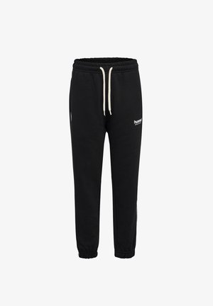 Młoda dziewczyna z warkoczami, ubrana w czarną i zieloną bluzę oraz czarne spodnie dresowe z białymi trampkami, patrzy w dół.