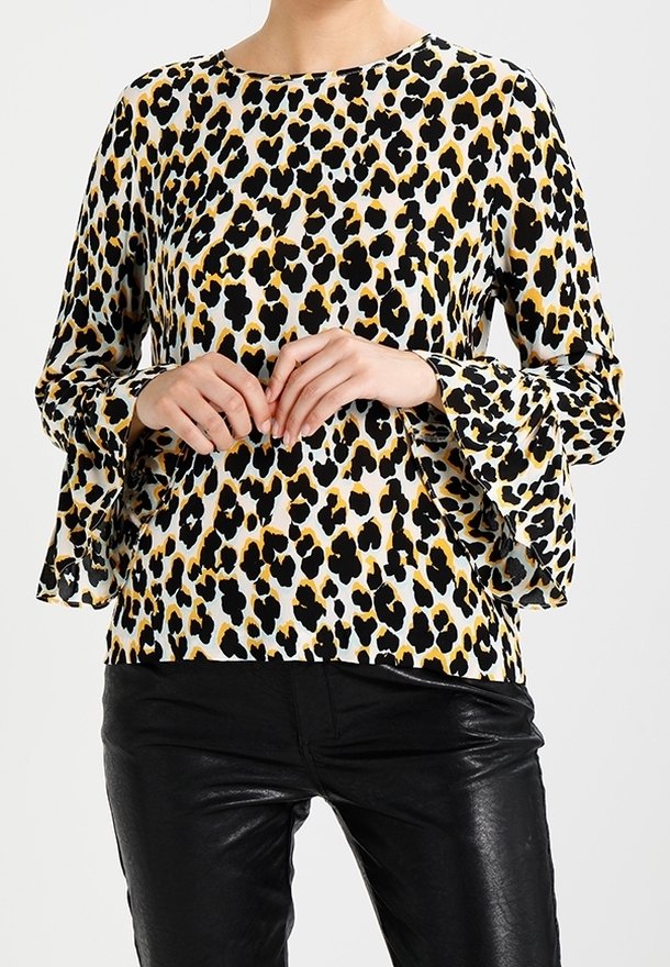 Blouse en imprimé léopard dans des tons beige, noir et or ; dotée de manches longues avec des poignets évasés. Portée avec des pantalons en cuir noir.