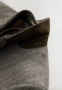 Brązowa tkanina w wzór jodełki z zagiętym kołnierzem. Wnętrze ma ciemną podszewkę. Staranne przeszycia wzdłuż krawędzi. Miękka faktura.