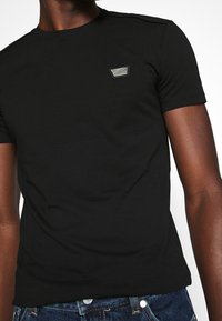 Schwarzes T-Shirt aus Baumwolle mit Rundhalsausschnitt, kurzen Ärmeln und einem kleinen, dreieckigen Logo-Patch auf der linken Brust.
