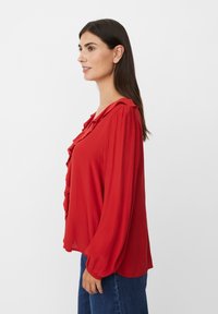 Rote Bluse mit gerafftem Ausschnitt, langen Ärmeln mit elastischen Manschetten, aus leichtem Stoff, getragen mit blauen Jeans. Seitenprofilansicht.