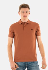 Bruine korte mouwen polo met kraag, voorzien van een klein zwart geborduurd logo op de linkerborst, in combinatie met blauwe spijkerbroeken.