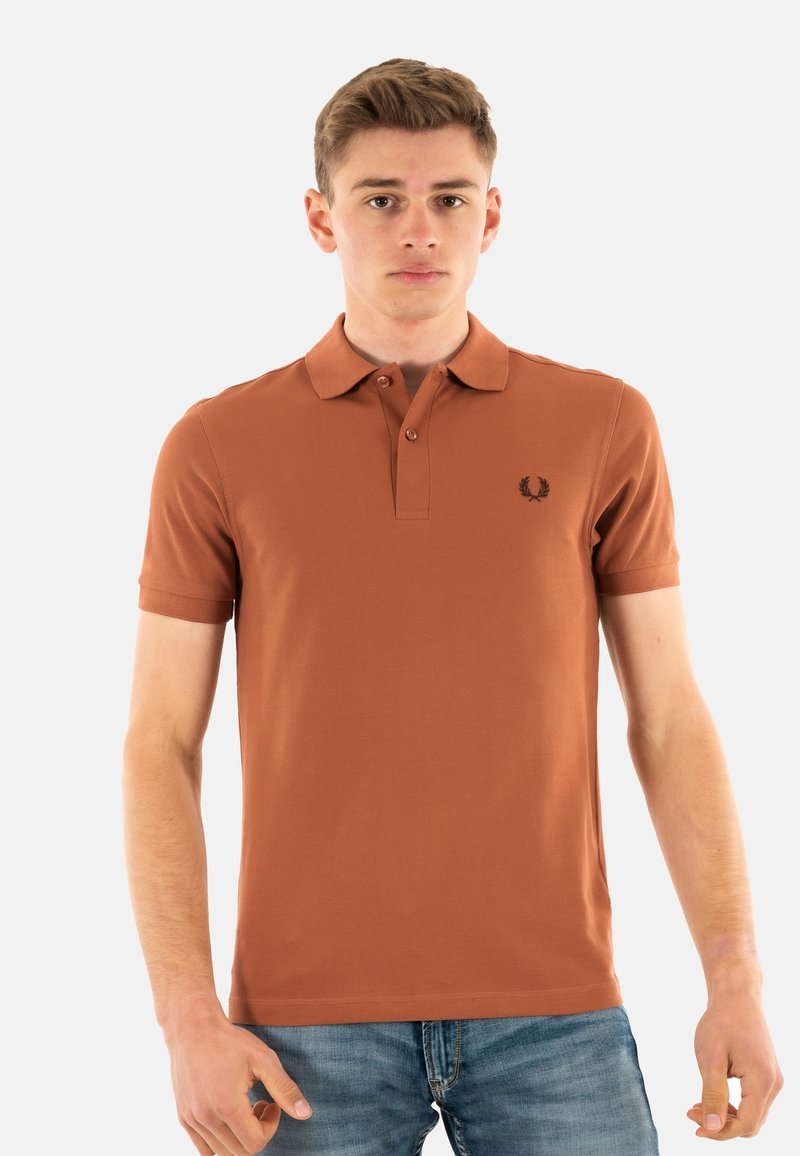 Bruine korte mouwen polo met kraag, voorzien van een klein zwart geborduurd logo op de linkerborst, in combinatie met blauwe spijkerbroeken.