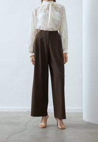 Blouse blanche texturée avec un nœud au cou ; associée à un pantalon large marron taille haute et des sandales dorées à lanières.
