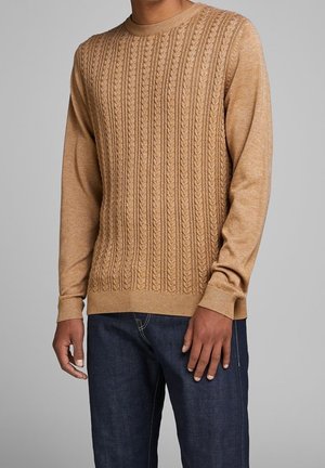 Man draagt een kabelgebreide trui in kamelkleur met lange mouwen en donkerblauwe jeans, staand tegen een effen grijze achtergrond.